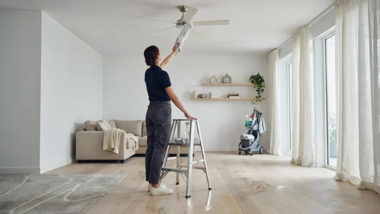 Professional cleaner dusting a ceiling fan in a bright modern living room using an extendable duster