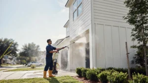 Professional technician pressure washing white vinyl siding of a two-story residential house on a sunny day.